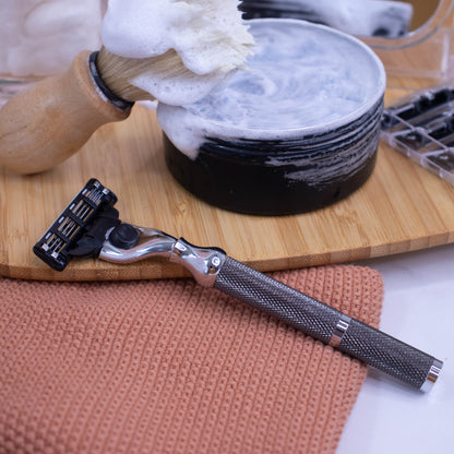 A dark silver-colored Parker cartridge razor handle with an all-metal textured grip displayed resting against a wooden tray with a close-up of the handle showing the chrome plating and a cartridge razor head.  Behind sits a tin of shaving soap with a shaving brush covered in the soap lather against a blurred bathroom background.