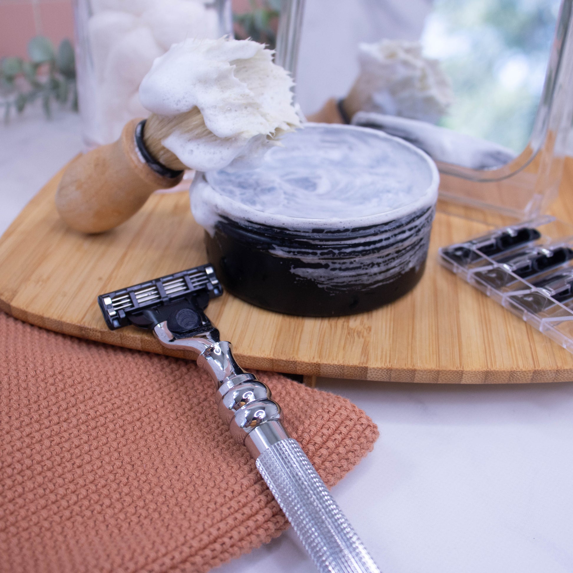 A silver-colored Parker cartridge razor handle with an all-metal textured grip displayed resting against a wooden tray with a close-up of the handle showing the chrome plating and a cartridge razor head.  Behind sits a tin of shaving soap with a shaving brush covered in the soap lather against a blurred bathroom background.