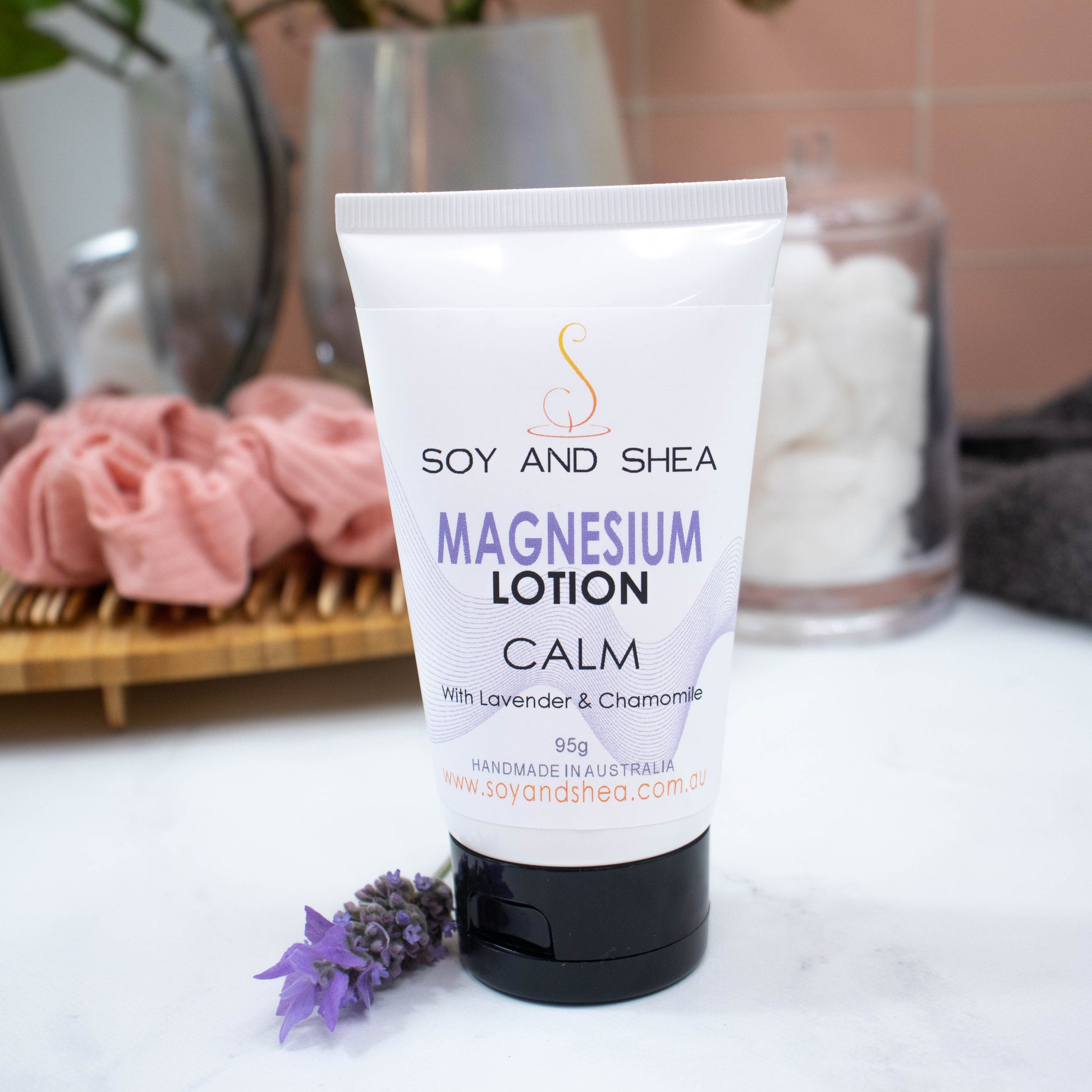 A tube of Soy and Shea's "Calm Magnesium Lotion" with Lavender and Chamomile essential oil rests on a white surface. The background features a wooden tray with pink scrunchies, a jar of cotton rounds, and soft towels. A sprig of purple flowers is placed near the cream.