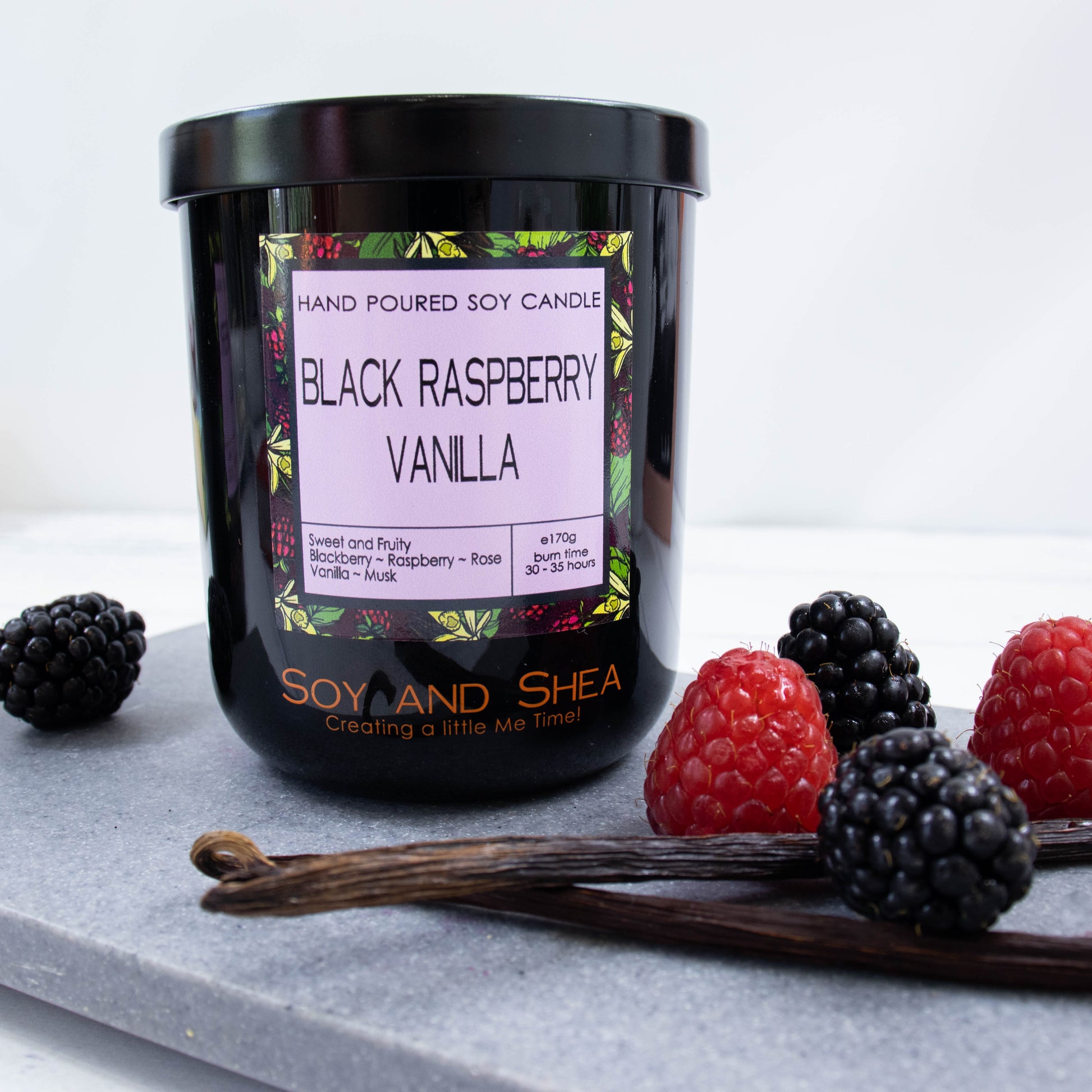 A black glass jar with black metal lid stands on the grey stone surface.  A decorative label is displayed on the front with information about the candle.   The candle is surrounded by a mix of black and red berries and two pods of vanilla.