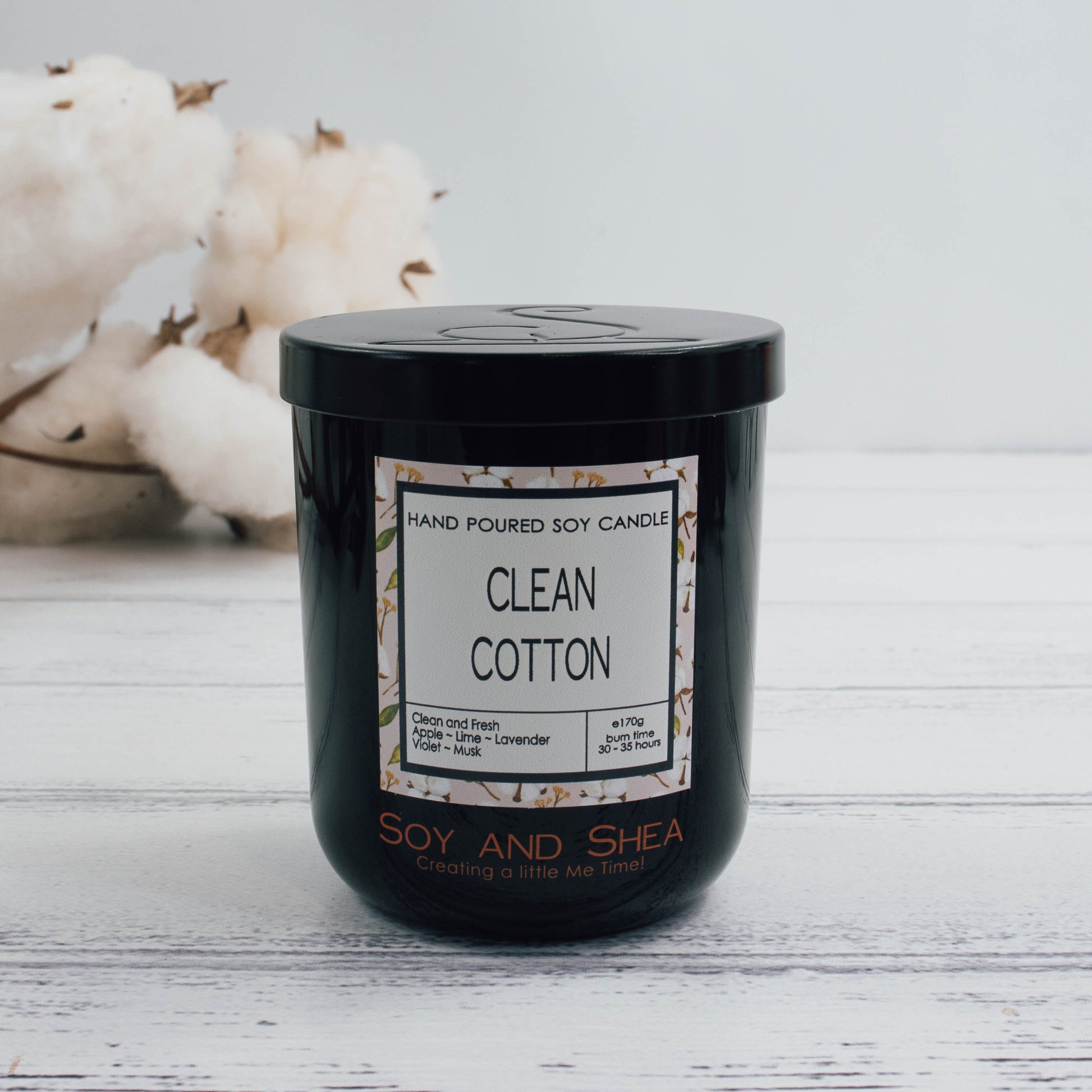 A black glass jar with black metal lid stands on the white wooden surface.  A decorative label is displayed on the front with information about the candle.   In the background is a branch of natural cotton bolls