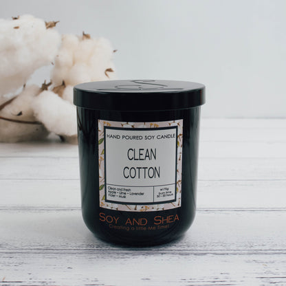 A black glass jar with black metal lid stands on the white wooden surface.  A decorative label is displayed on the front with information about the candle.   In the background is a branch of natural cotton bolls
