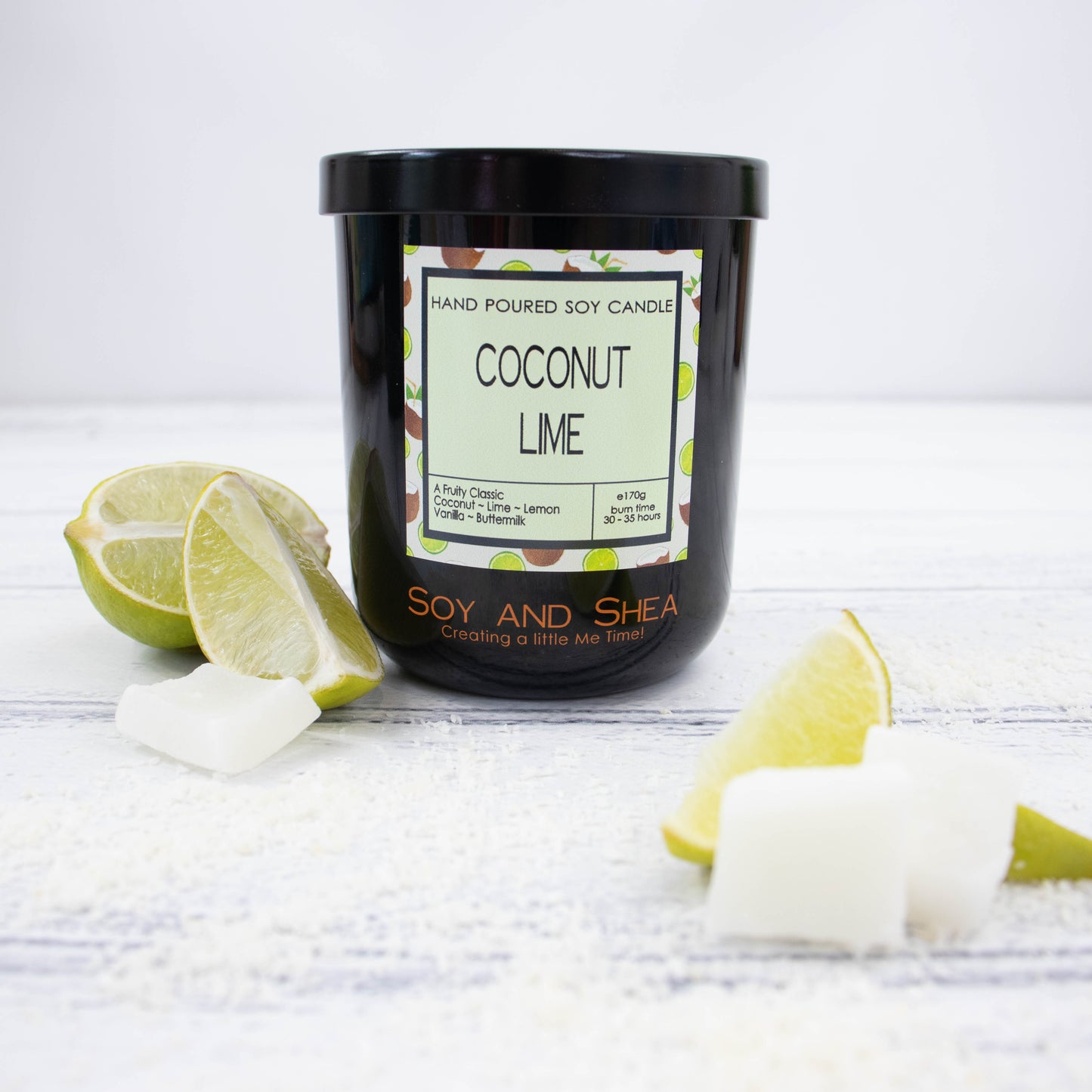 A black glass jar with black metal lid stands on the wooden side.  A decorative label is displayed on the front with information about the candle.   Surroundleing the candle are wedges of lime and piece of coconut flesh.