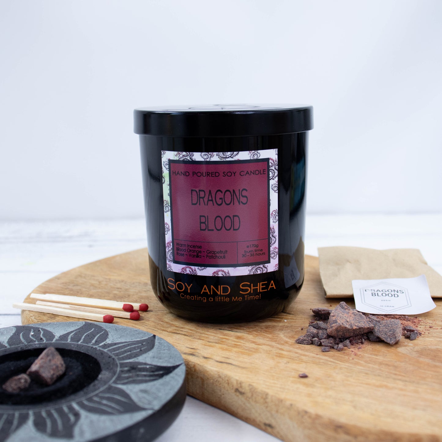 A black glass jar with black metal lid sits on the wooden side.  A decorative label is displayed on the front with information about the candle.  Surrounding the candle are pieces of resin with a paper bag labelled DRAGONS BLOOD along with a resin burner and matches.