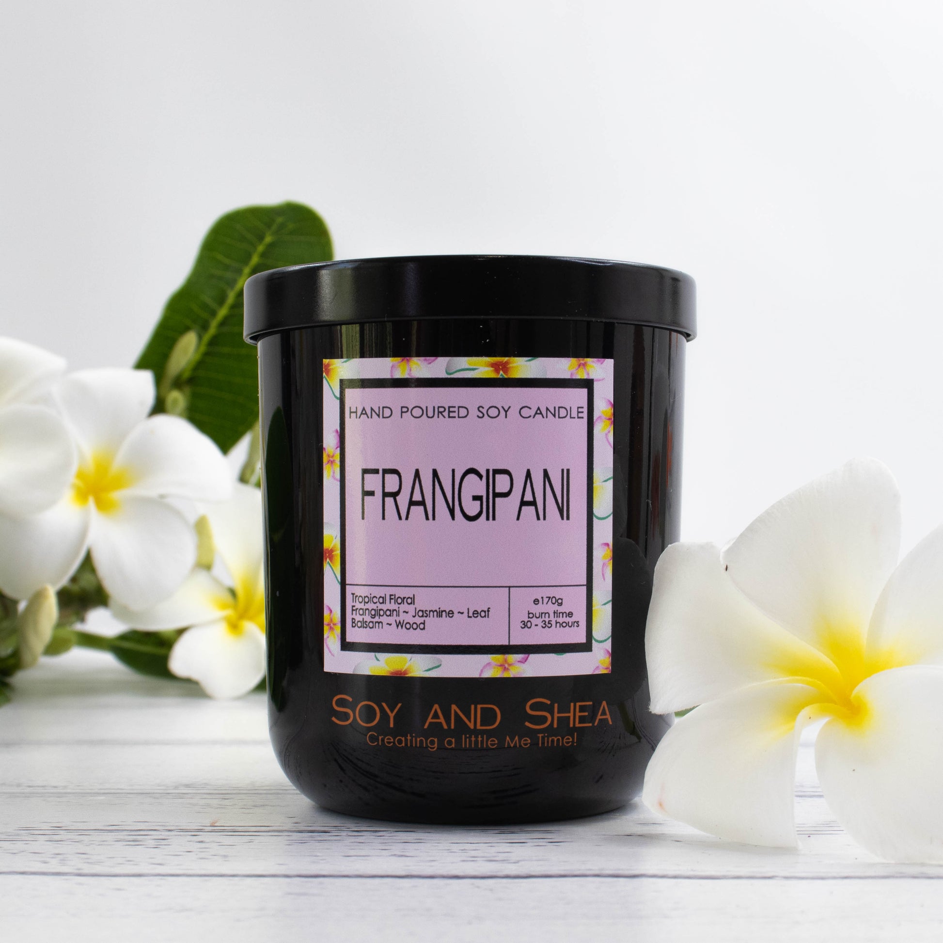 A black glass jar with black metal lid stands on the white wooden surface.  A decorative label is displayed on the front with information about the candle.   The candle is surrounded white franganpis with yelllow centres.