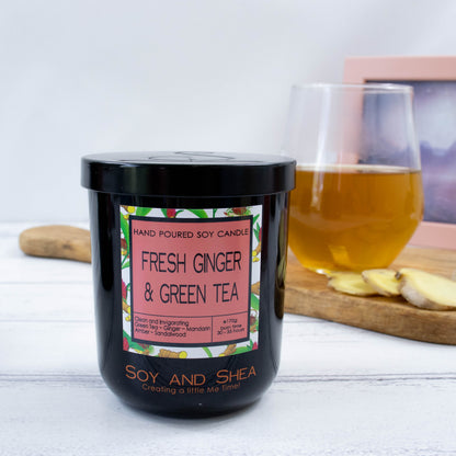A black glass jar with black metal lid stands on the wooden side.  A decorative label is displayed on the front with information about the candle.   Behind the candle on a wooden tray is a a clear glass of tea along with some slices of ginger.