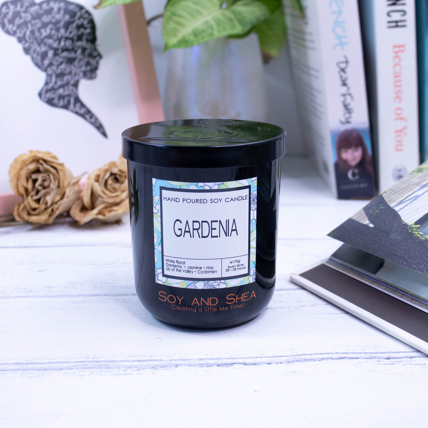 A black glass jar with black metal lid sits on the wooden side.  A decorative label is displayed on the front with information about the candle.  In the background is a picture and some books.