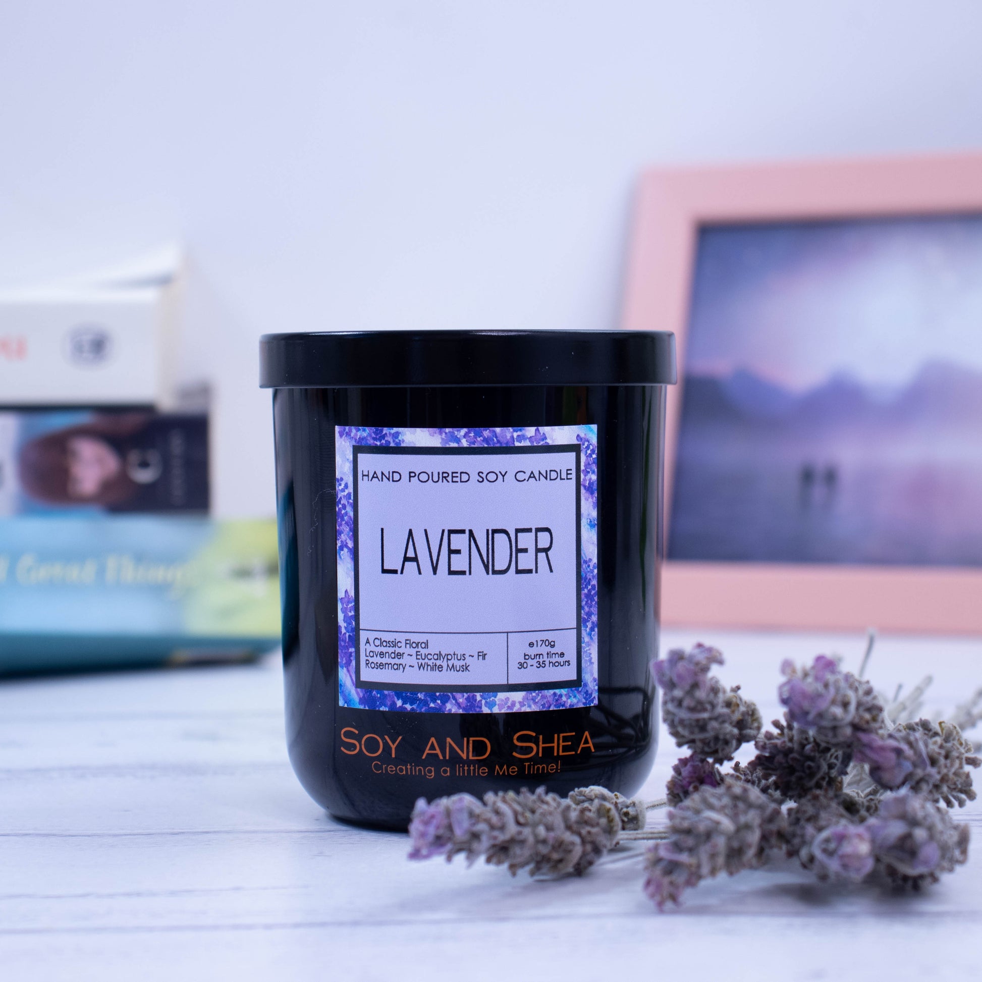 A black glass jar with black metal lid sits on the wooden side.  A decorative label is displayed on the front with information about the candle.  The candle is surrounded by dry lavender.  In the background is a picture and some books.
