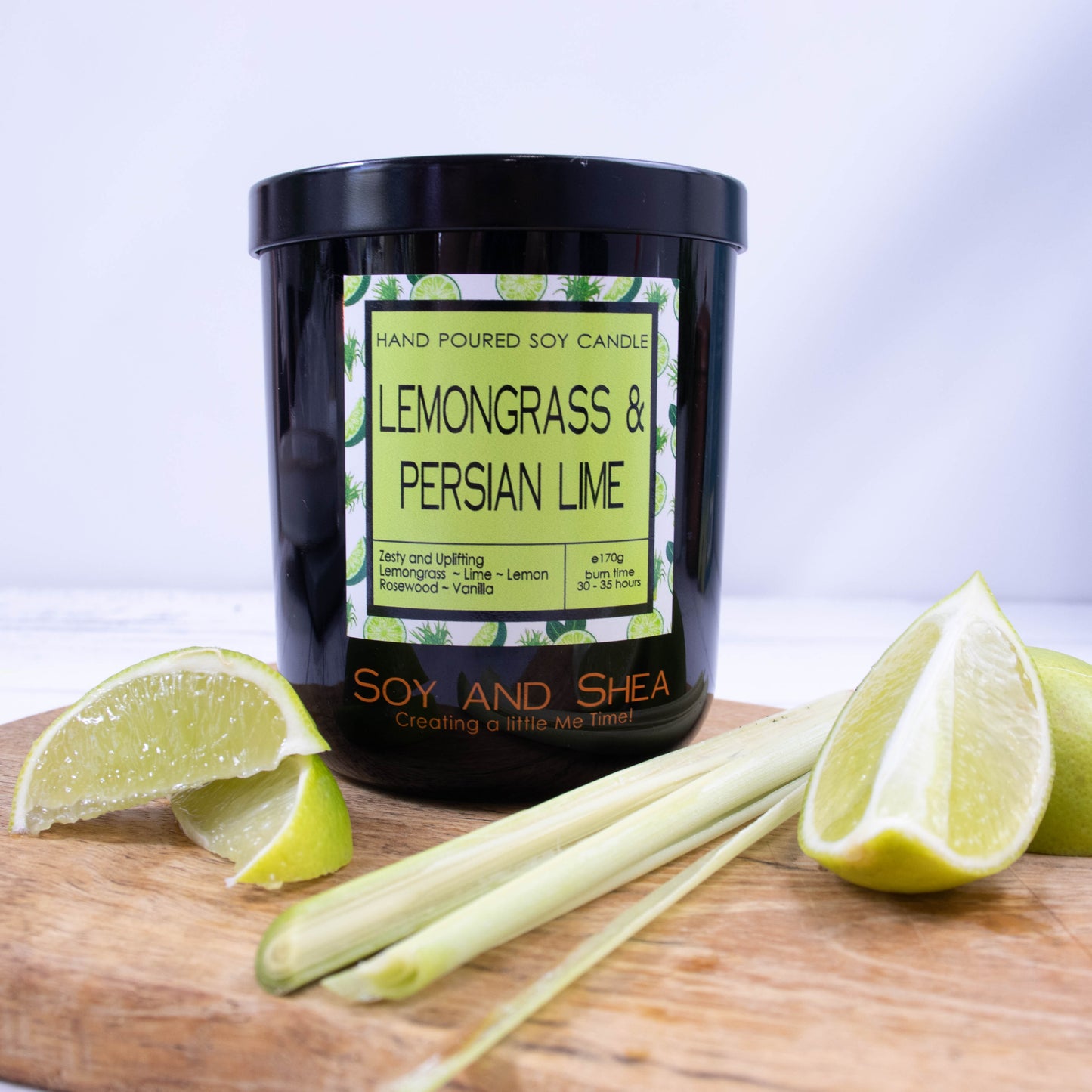A black glass jar with black metal lid stands on the wooden side.  A decorative label is displayed on the front with information about the candle.   Beside the candle are some slices of lime and a stalk of lemongrass.