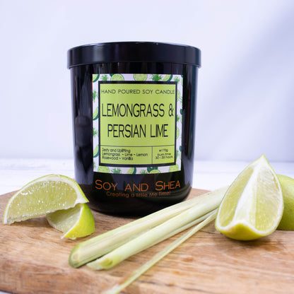 A black glass jar with black metal lid stands on the wooden side.  A decorative label is displayed on the front with information about the candle.   Beside the candle are some slices of lime and a stalk of lemongrass.