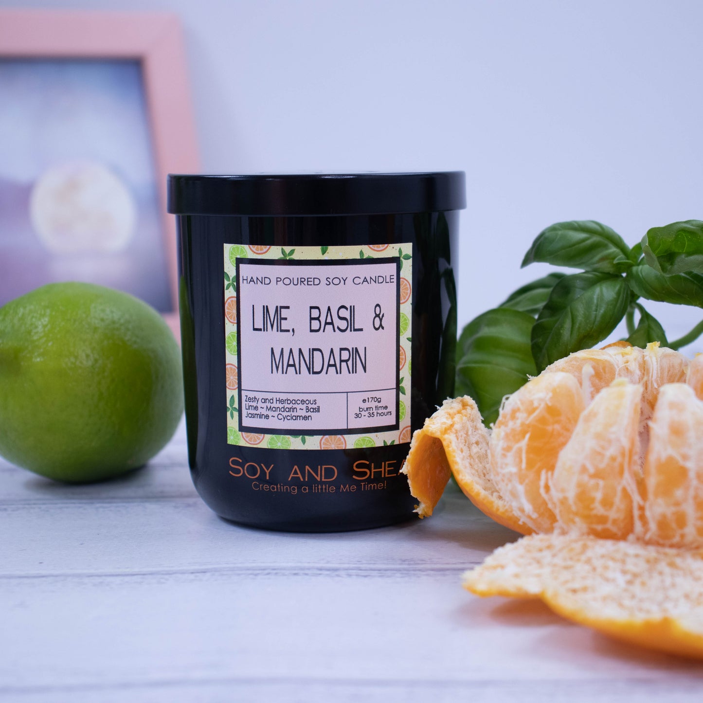 A black glass jar with black metal lid sits on the wooden side.  A decorative label is displayed on the front with information about the candle. The candle is surronded by a peeled mandarin, whole lime and basil leaves.