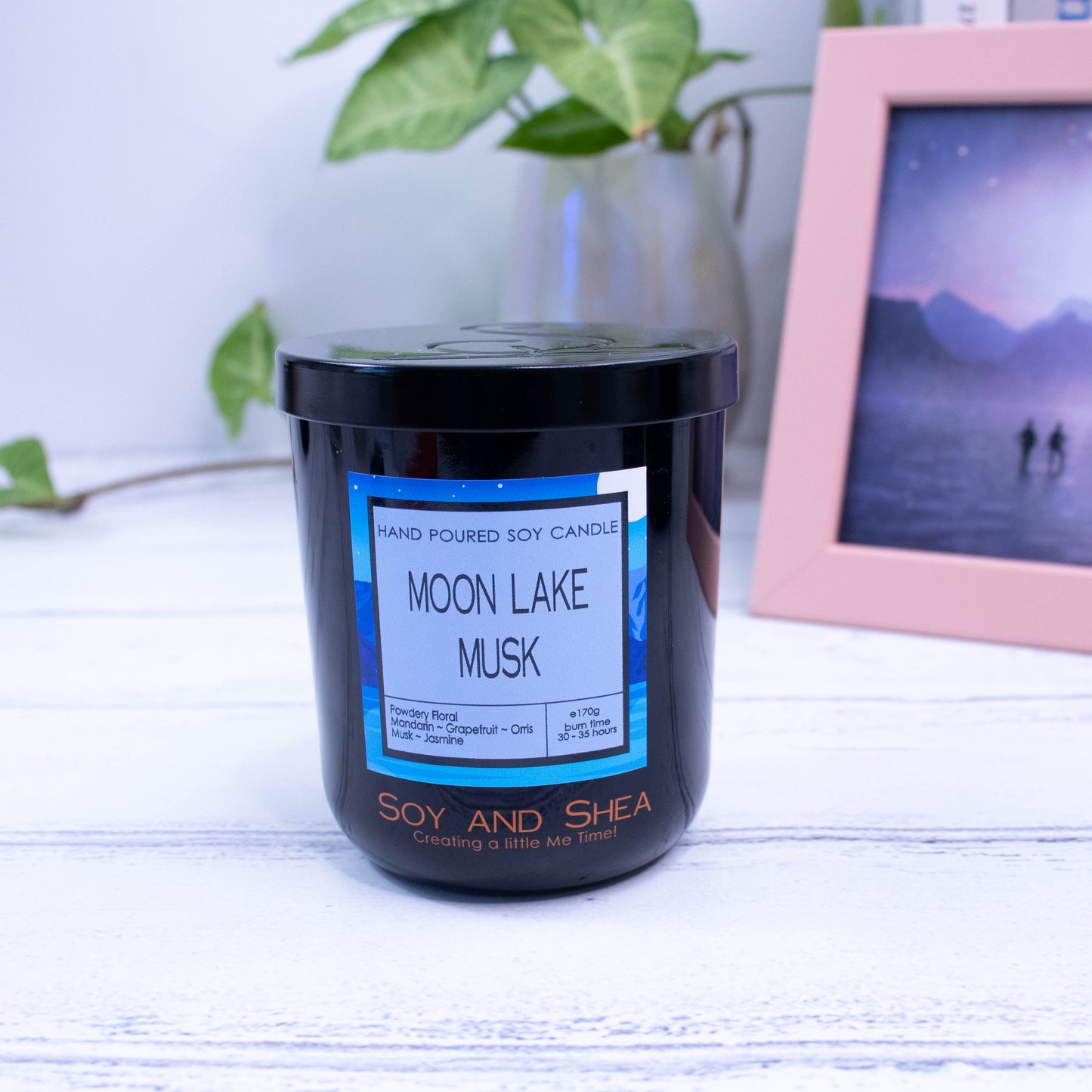 A black glass jar with black metal lid sits on the wooden side.  A decorative label is displayed on the front with information about the candle.  In the background is a picture and a vase with a green plant.