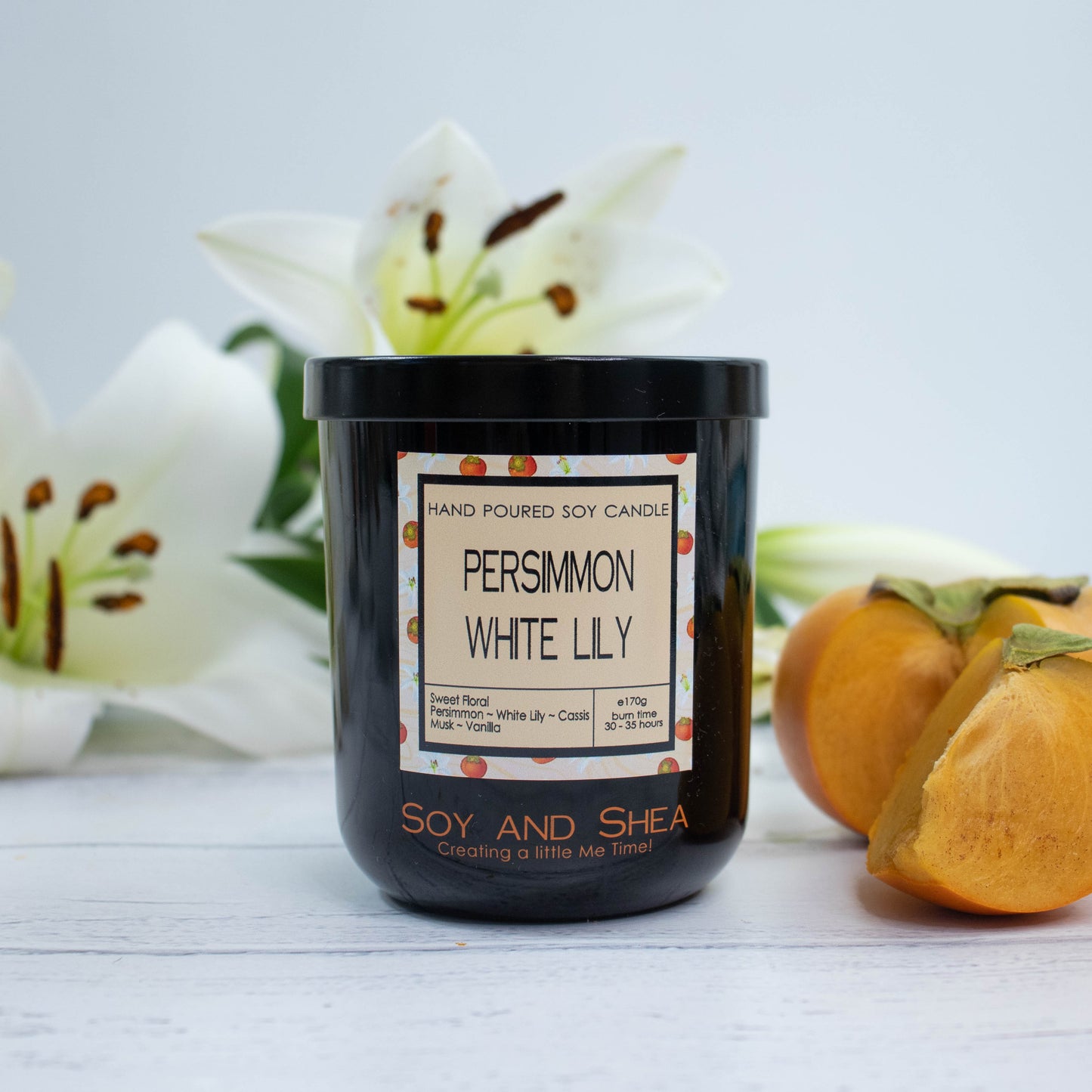 A black glass jar with black metal lid sits on the wooden side.  A decorative label is displayed on the front with information about the candle.  To the right of the candle is a ripe persimmon with a wedge cut out and behind are two white lily flowers.