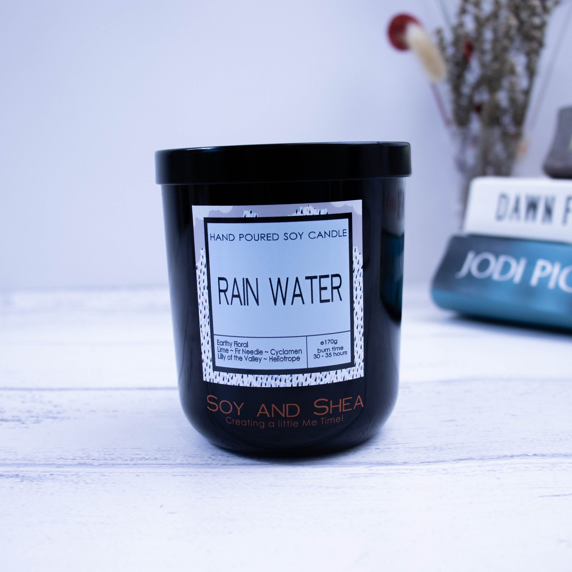 A black glass jar with black metal lid sits on the wooden side.  A decorative label is displayed on the front with information about the candle.  In the background is a stack of books besidea vase with dried flowers.