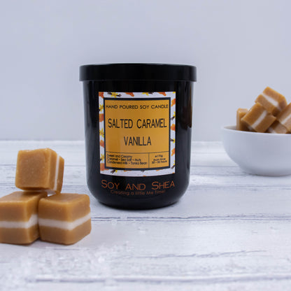 A black glass jar with black metal lid sits on the wooden side.  A decorative label is displayed on the front with information about the candle.  To the left of the candle is a three caramel lollies with a small pile of salt.  In the background is white bowl filled with caramels.