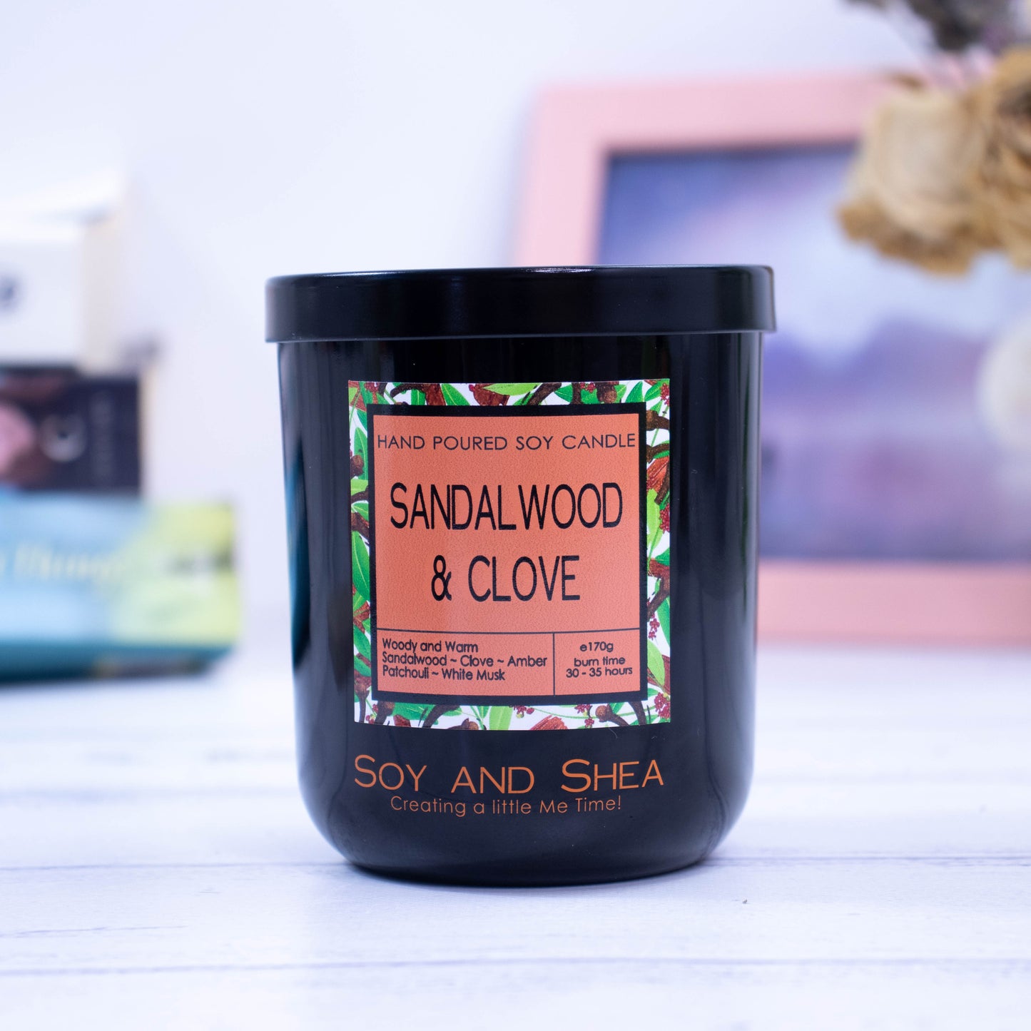 A black glass jar with black metal lid sits on the wooden side.  A decorative label is displayed on the front with information about the candle.  In the background is a picture and some books.