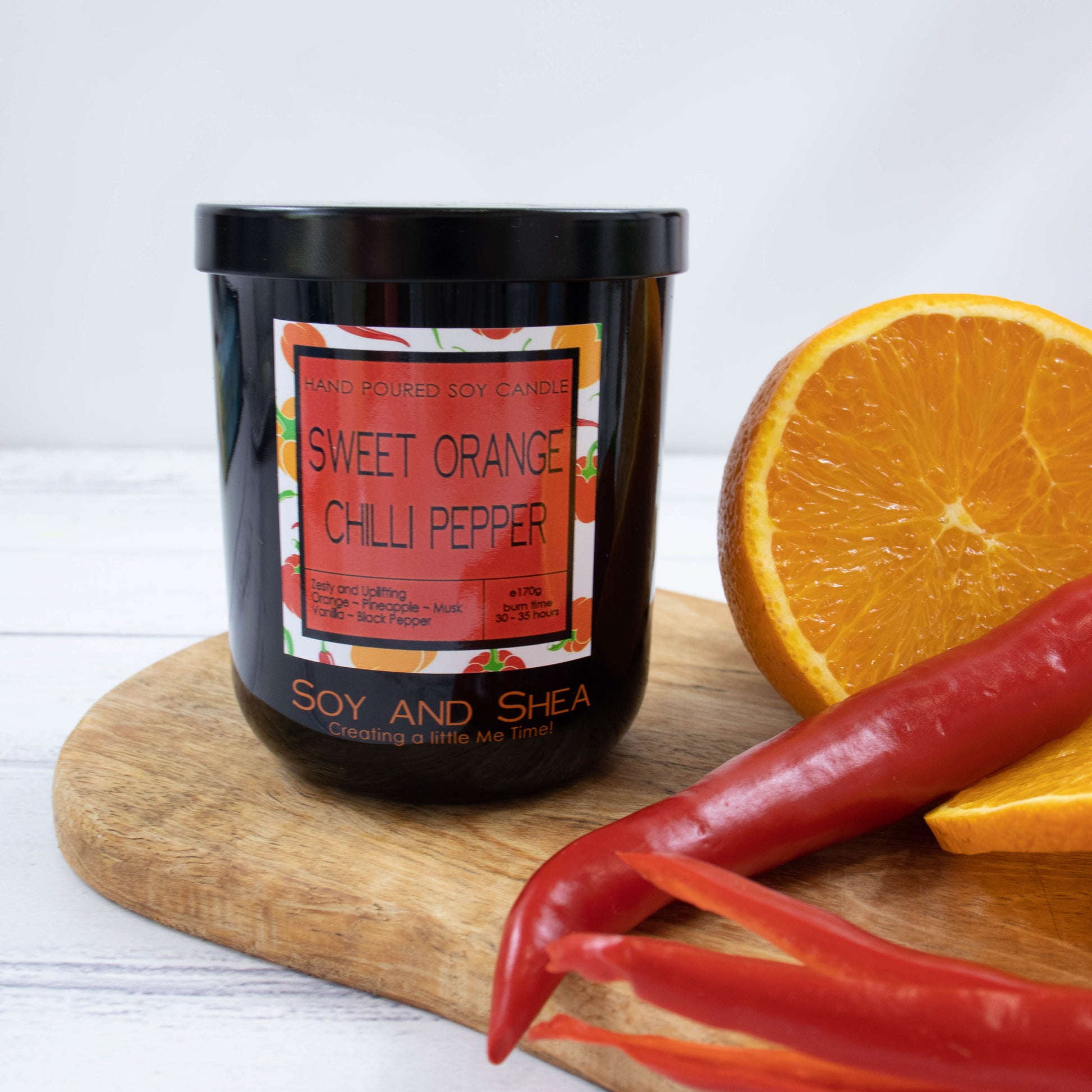 A black glass jar with black metal lid stands on the wooden side.  A decorative label is displayed on the front with information about the candle.   Beside the candle sits an orange that has been cut in half and some red chillies.