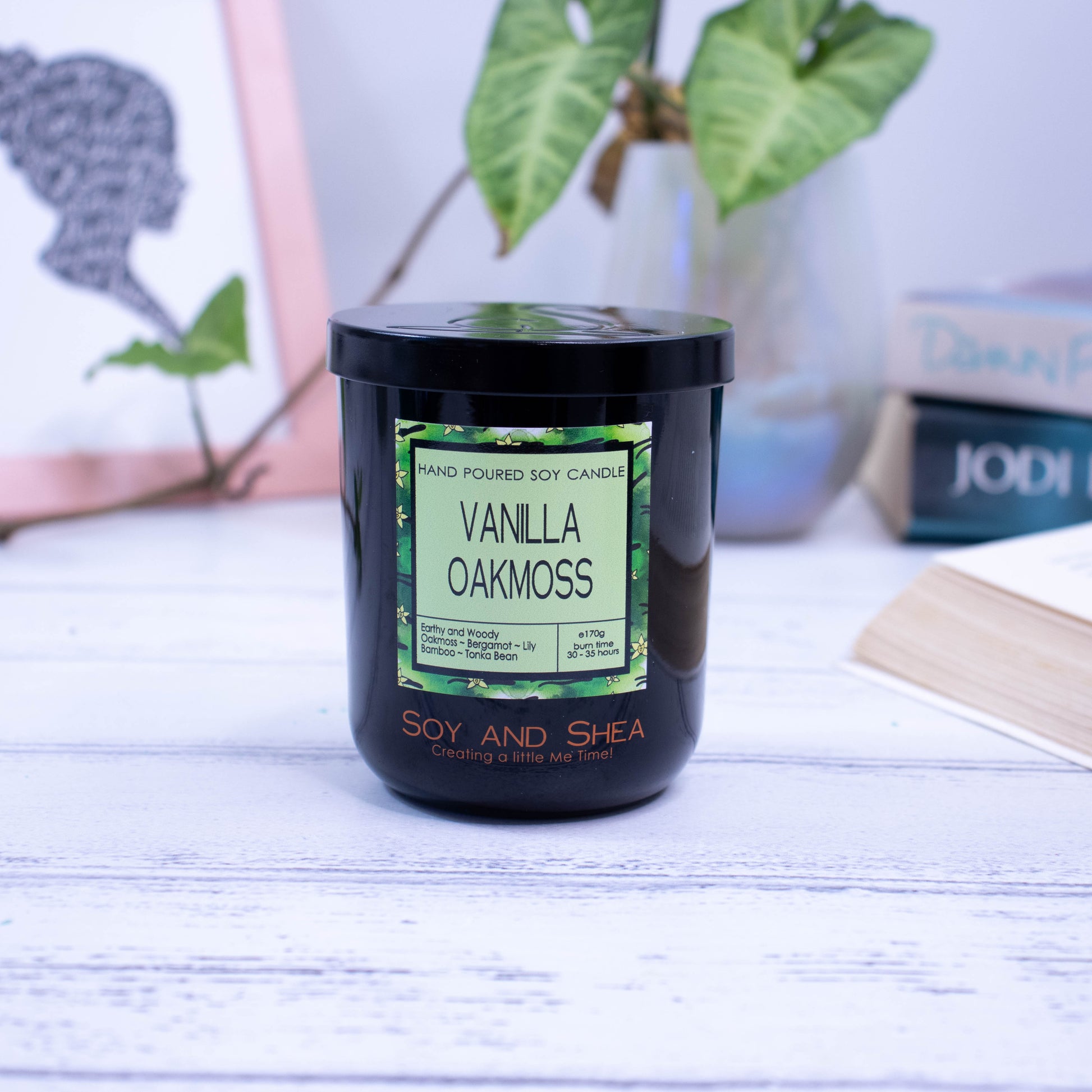 A black glass jar with black metal lid sits on the wooden side.  A decorative label is displayed on the front with information about the candle.  In the background is a picture and some books.