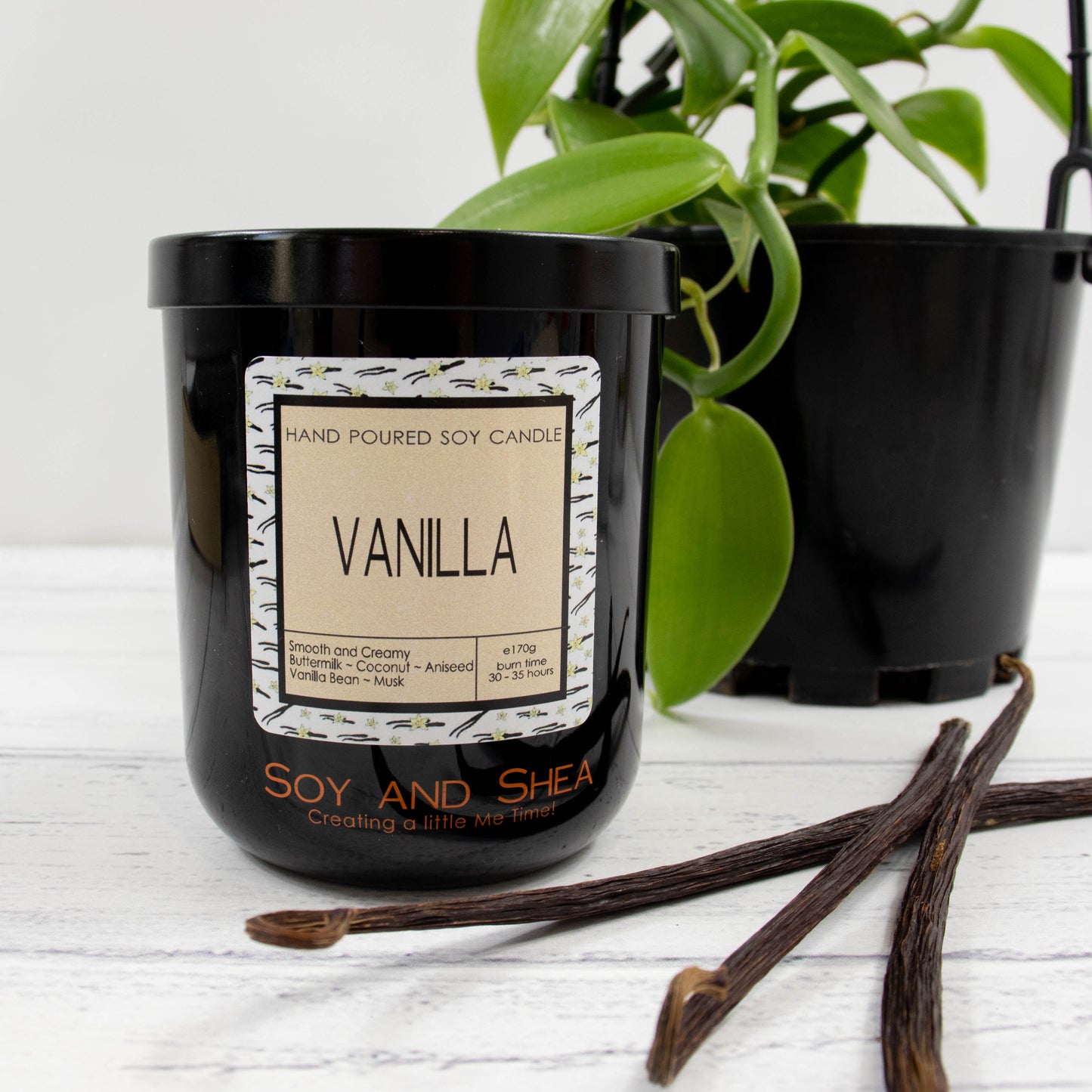 A black glass jar with black metal lid stands on the wooden side.  A decorative label is displayed on the front with information about the candle.   Beside the candle lies three dried vanilla  pods and a potted vanilla vine can be seen in the background.