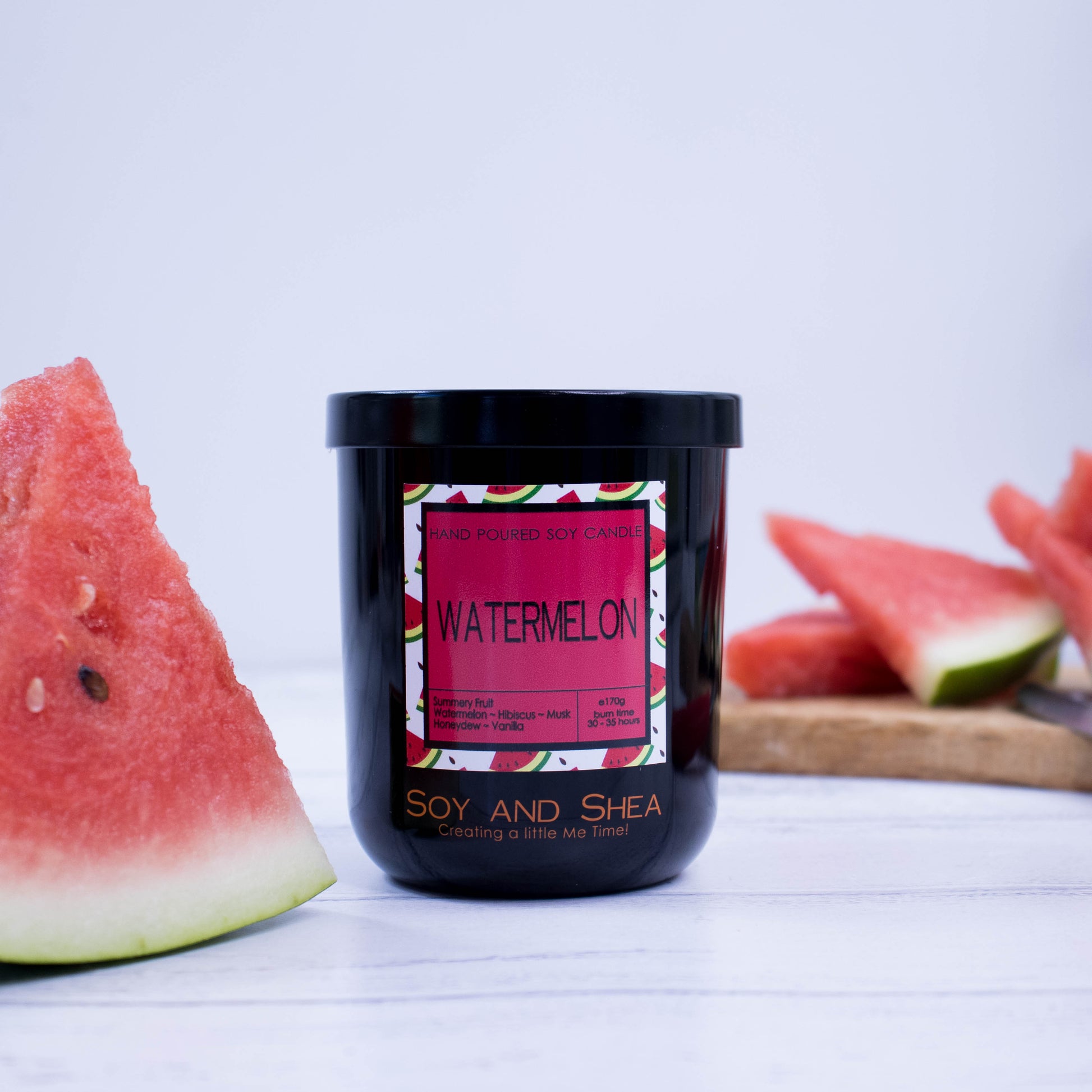A black glass jar with black metal lid sits on the wooden side.  A decorative label is displayed on the front with information about the candle.  To the left of the candle is a piece of watermelon standing upright and in the blurred background is a wooden tray with more watermelon slices.