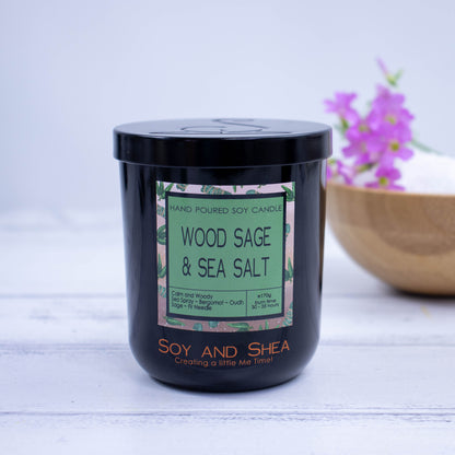 A Black jar with colourful product label and black metal lid stands on a wooden surgface with bright white background.  A wooden bowl with purple flowers sit behind the candle.