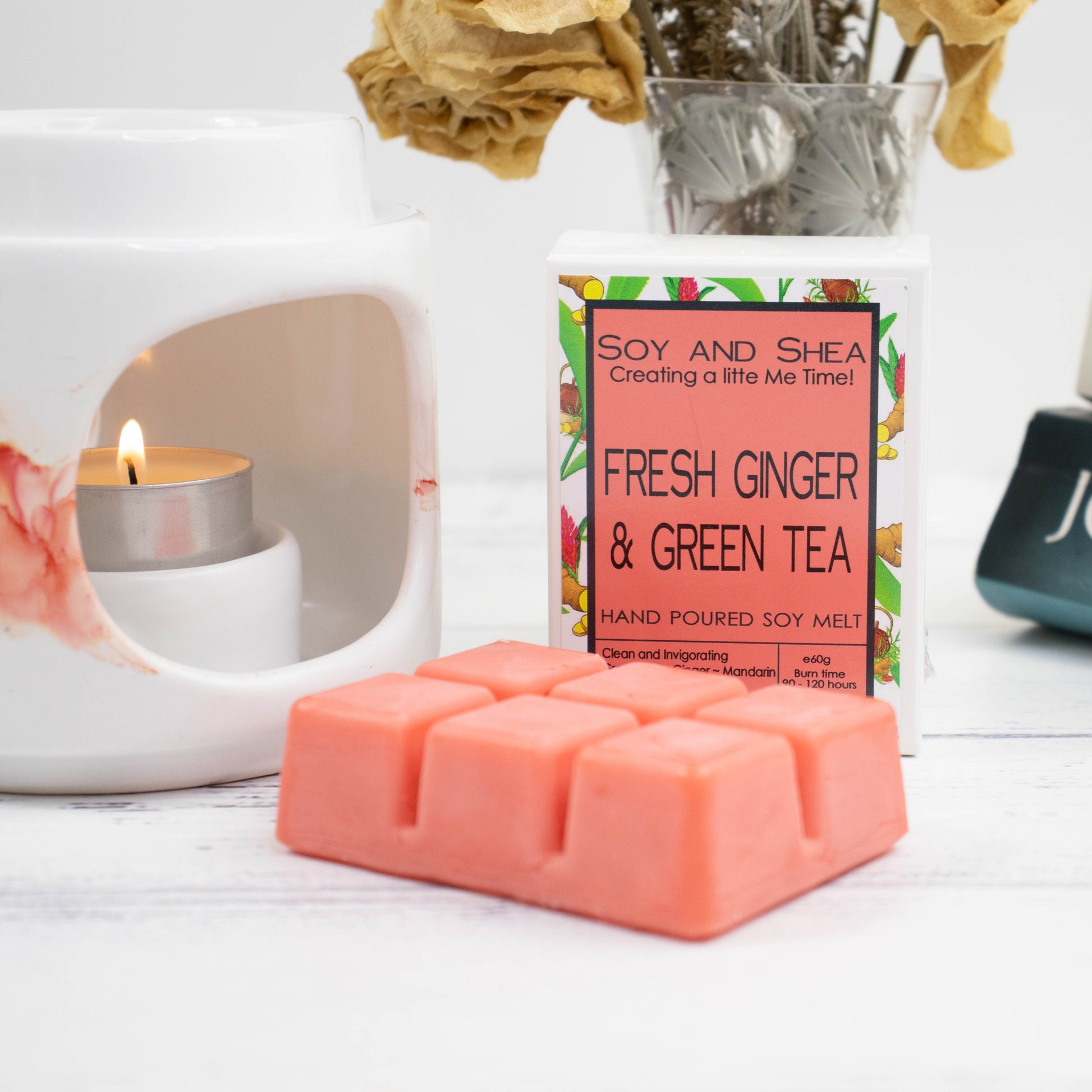 A lit white ceramic burner sits next to a package of Soy and Shea's clearance "Soy Melt Blocks" in the "Fresh Ginger & Green Tea" scent. In front of the package is a pink segmented wax melt, while dried flowers and a glass jar in the background contribute to the eco-friendly home fragrance ambiance.