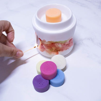 A hand holds a match to light a fragrance warmer.  The warmer is white with orange and gold marbling around the middle.  An orange melt puck sits in the top of the fragrance warms with a stack of melt pucks in various colours including purple, pink and blue sitting in front of the warmer. 