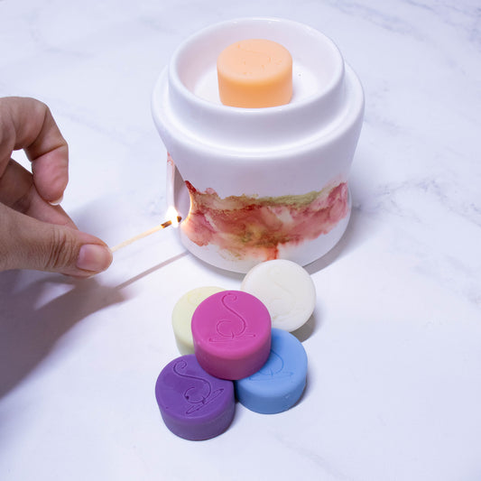 A hand holds a match to light a fragrance warmer.  The warmer is white with orange and gold marbling around the middle.  An orange melt puck sits in the top of the fragrance warms with a stack of melt pucks in various colours including purple, pink and blue sitting in front of the warmer. 