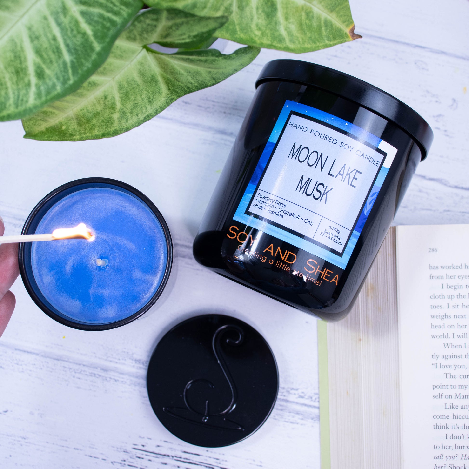 A flat lay photo showing a large black jar lying on its side on a white wooden surface next to a book and plant.  To the bottom of the jar is a smaller candle standing upright and filled with bright blue wax.  A hand reaches in with a lit match.