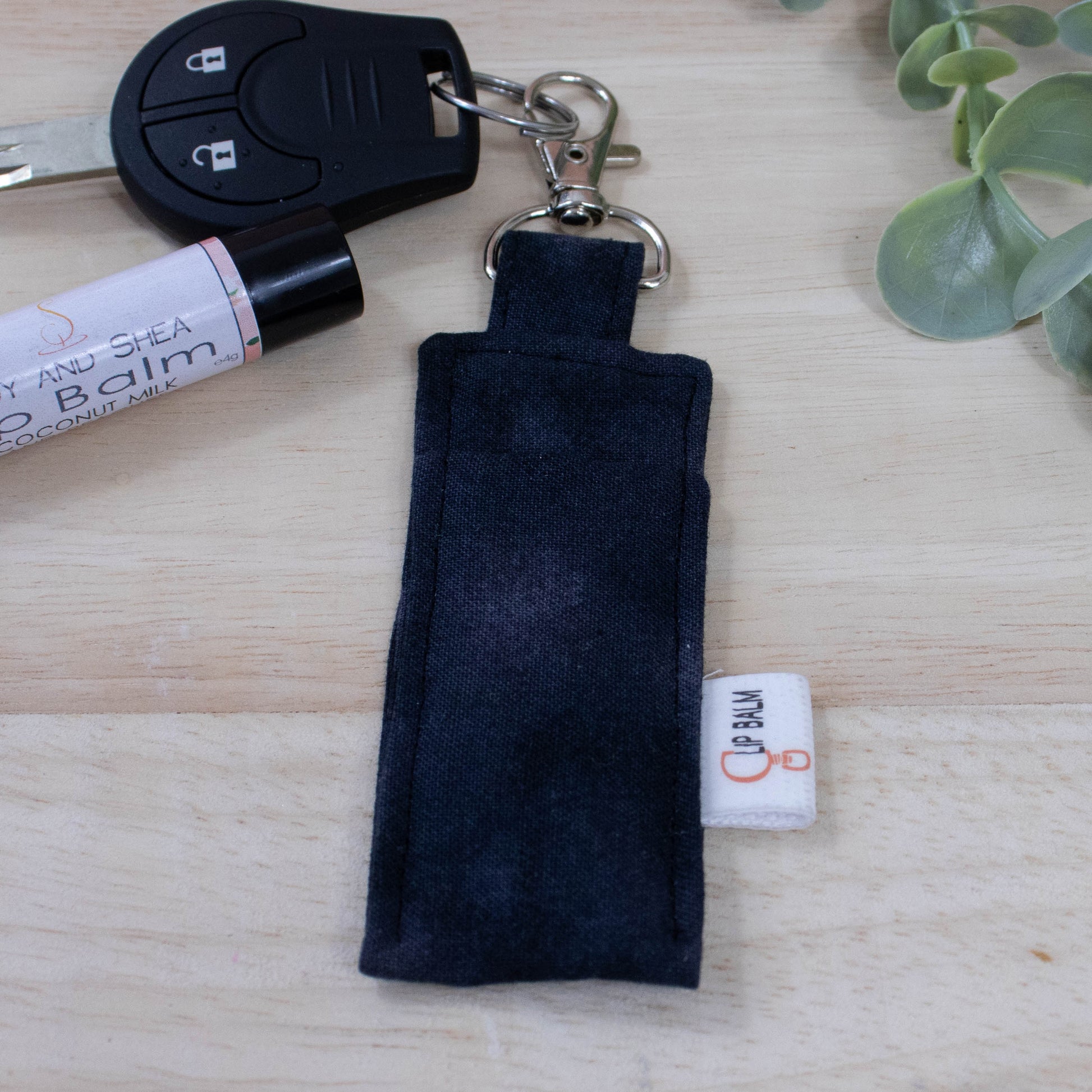 A clip balm holder lies on a wooden surface attached to a car key with a "lobster claw" clip.  To the side is a tube of lip balm and some greenery.  The Clip Balm is made from a mottled black fabric.