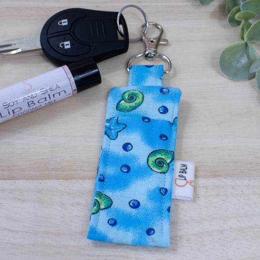 A clip balm holder lies on a wooden surface attached to a car key with a "lobster claw" clip.  To the side is a tube of lip balm and some greenery.  The Clip Balm has a bright blue mottled background with illustrations of shells, starfish and bubbles randomly placed.
