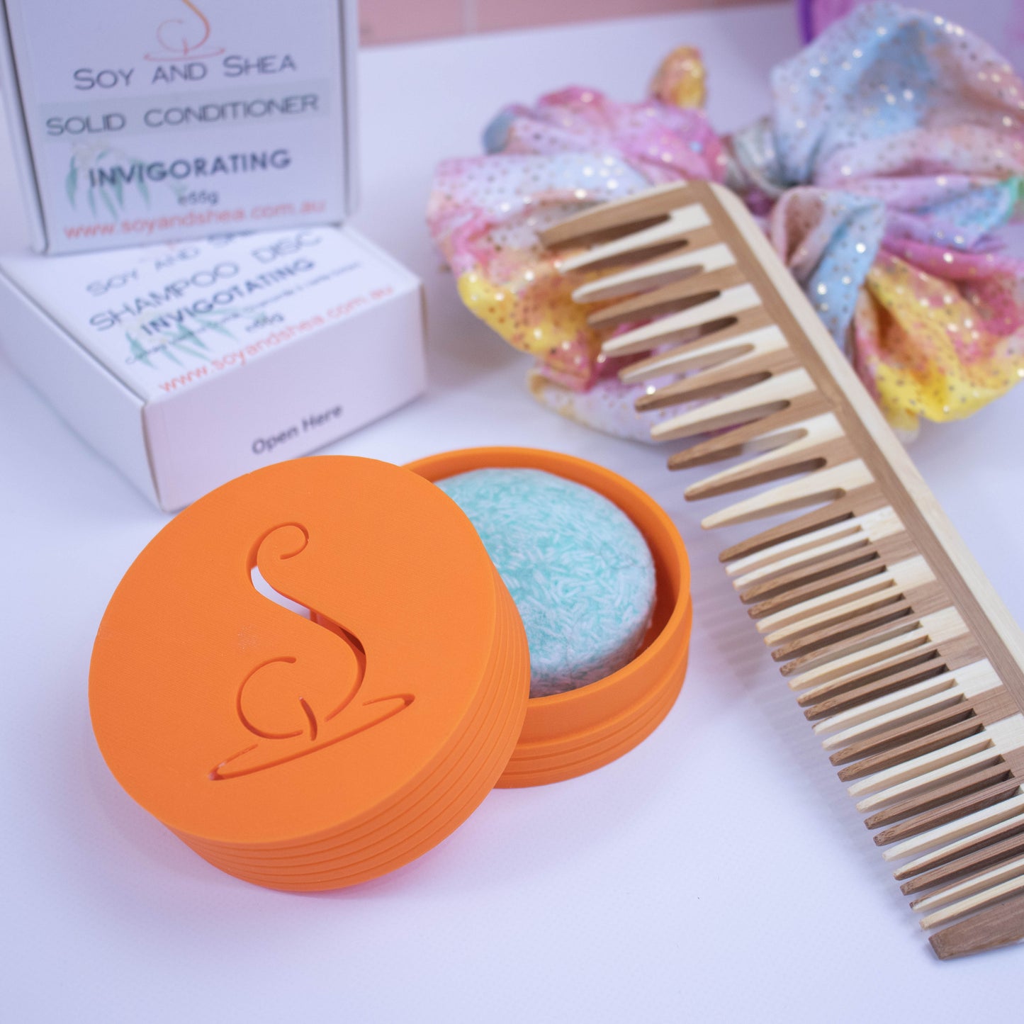 An orange cylindrical pot with the Soy and Shea logomark cut out in the top, sits on a white surface.  The lid has been removed and is resting on the side of the base which holds a solid shampoo bar.  In the background stands the shampoo packaging next to a wooden comb and colourful scrunchie with shiny gold dots.