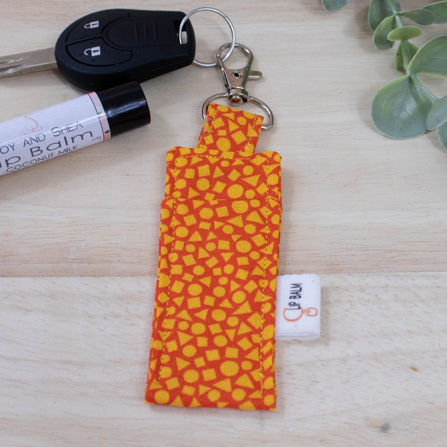 A clip balm holder lies on a wooden surface attached to a car key with a "lobster claw" clip.  To the side is a tube of lip balm and some greenery.  The Clip Balm features a random pattern of small yellow triangle, squares and circles against a bright orange background