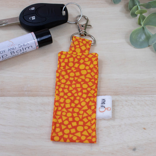 A clip balm holder lies on a wooden surface attached to a car key with a "lobster claw" clip.  To the side is a tube of lip balm and some greenery.  The Clip Balm features a random pattern of small yellow triangle, squares and circles against a bright orange background