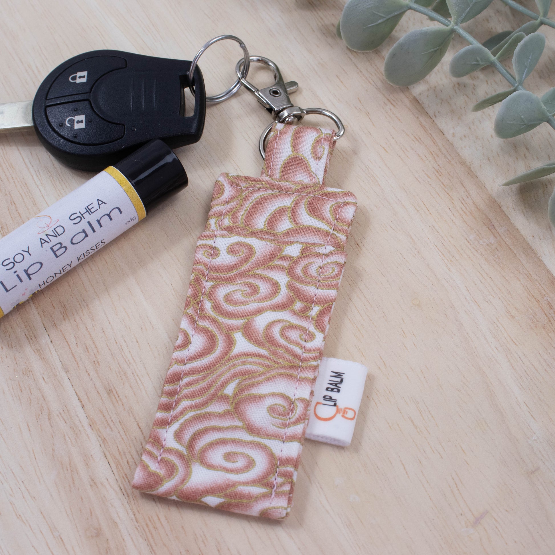 A clip balm holder lies on a wooden surface attached to a car key with a "lobster claw" clip.  To the side is a tube of lip balm and some greenery.  The Clip Balm has a white background covered in stylised clouds in peach and gold with an oriental feel.
