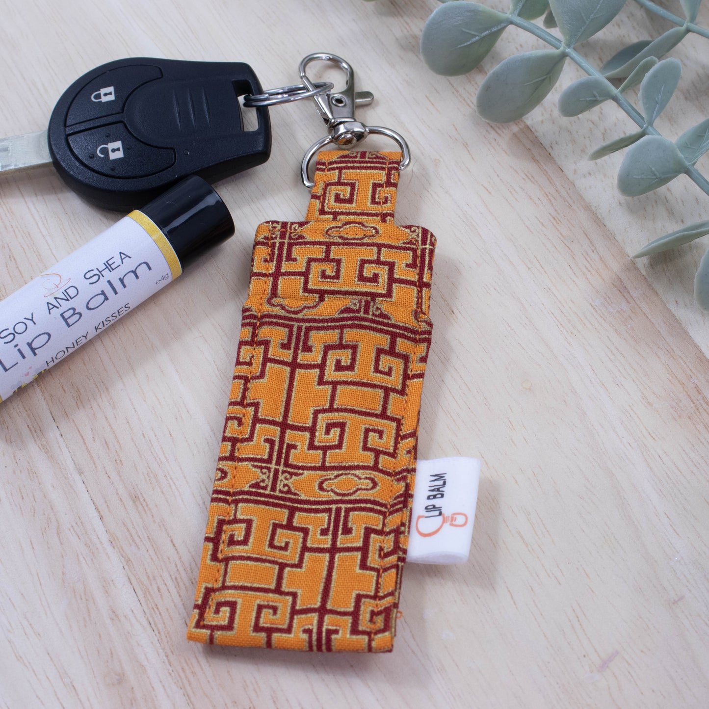 A clip balm holder lies on a wooden surface attached to a car key with a "lobster claw" clip.  To the side is a tube of lip balm and some greenery.  The Clip Balm has a bright orange background with a deep red line pattern with gold outlines that reflects oriental design.