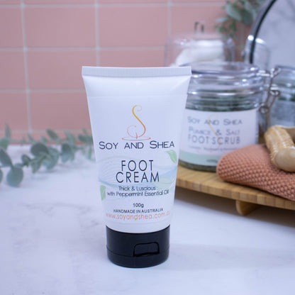 A tube of Soy and Shea foot cream placed on a bathroom countertop with a blurred background featuring pink tiles, greenery, and bathroom essentials.  The white tube with black flip-top lid, features a label that has a light blue wavy design with watercolour green leaves dotted around.