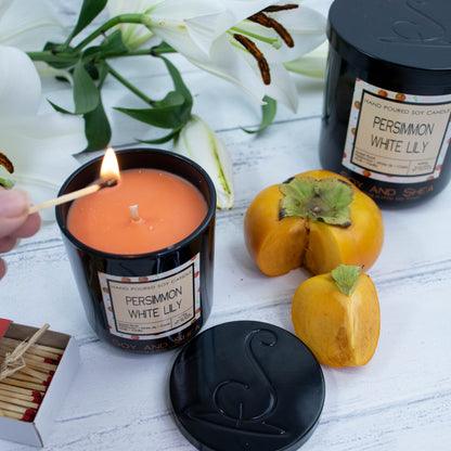A medium black glass candle stands on a white wooden surface with the lid removed to show an orange wax.  A hand reaches in with a lit match.  The lid of the candle is black metal and debossed with the logomark.  Surround the candle is a spray of white lillies along with a ripe persimmon and a larger candle to the back.