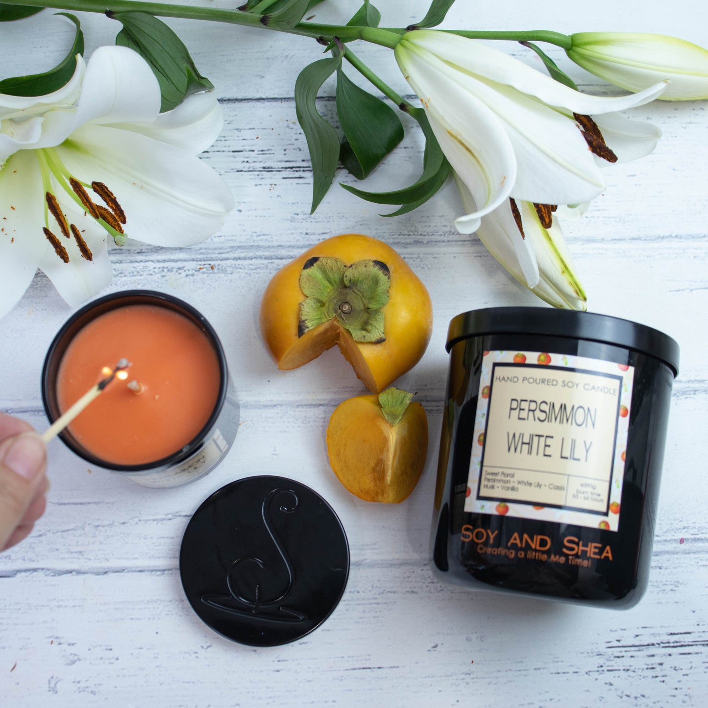 A flat lay photo showing a large black jar lying on its side on a white wooden surface with a spray of white lillies and a persimmon with a wedge cut out.  To the left of the jar is a smaller candle standing upright and filled with dark orange wax.  A hand reaches in with a lit match.