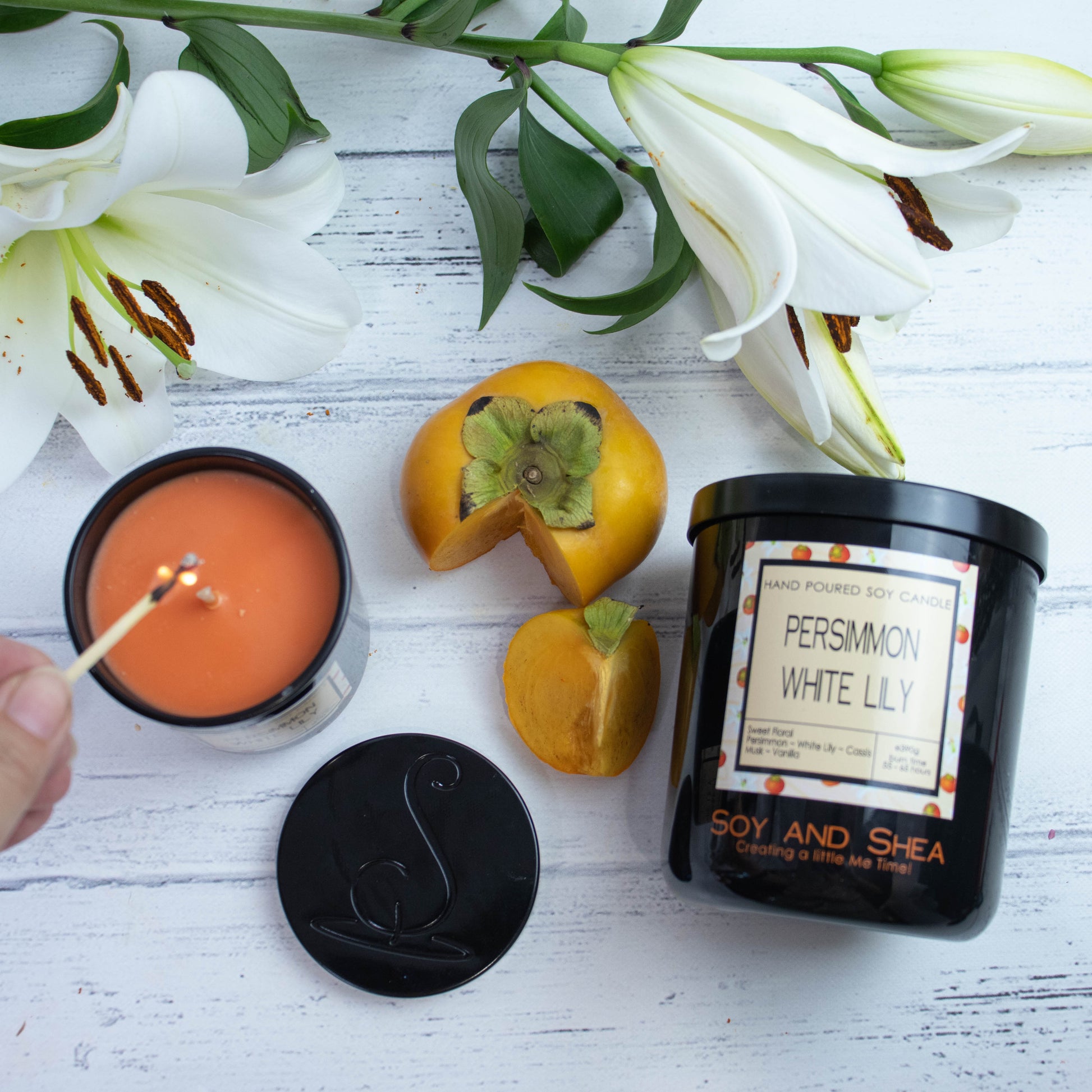 A flat lay photo showing a large black jar lying on its side on a white wooden surface with a spray of white lillies and a persimmon with a wedge cut out.  To the left of the jar is a smaller candle standing upright and filled with dark orange wax.  A hand reaches in with a lit match.