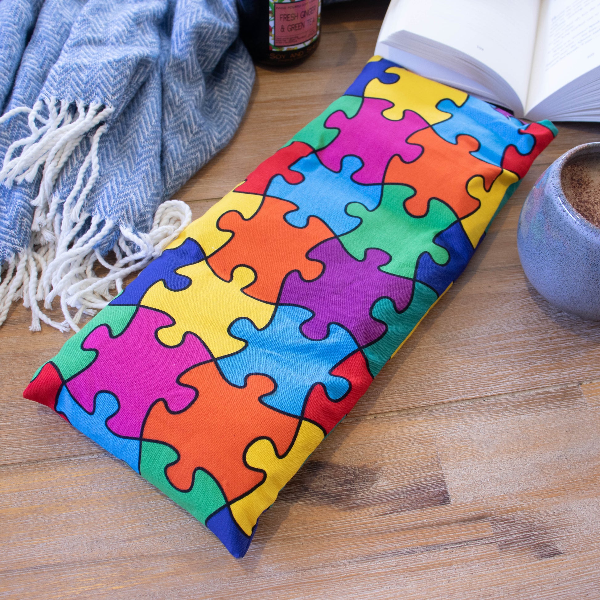 A long rectangular heat pack lies on a wooden surface surrounded by a blue blanket, open book, burning candle and filled coffee cup.  The Heat Pack shows connected jigsaw pieces in bright colours of pink, blue, purple orange, red and yellow.