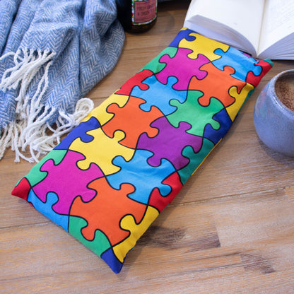 A long rectangular heat pack lies on a wooden surface surrounded by a blue blanket, open book, burning candle and filled coffee cup.  The Heat Pack shows connected jigsaw pieces in bright colours of pink, blue, purple orange, red and yellow.
