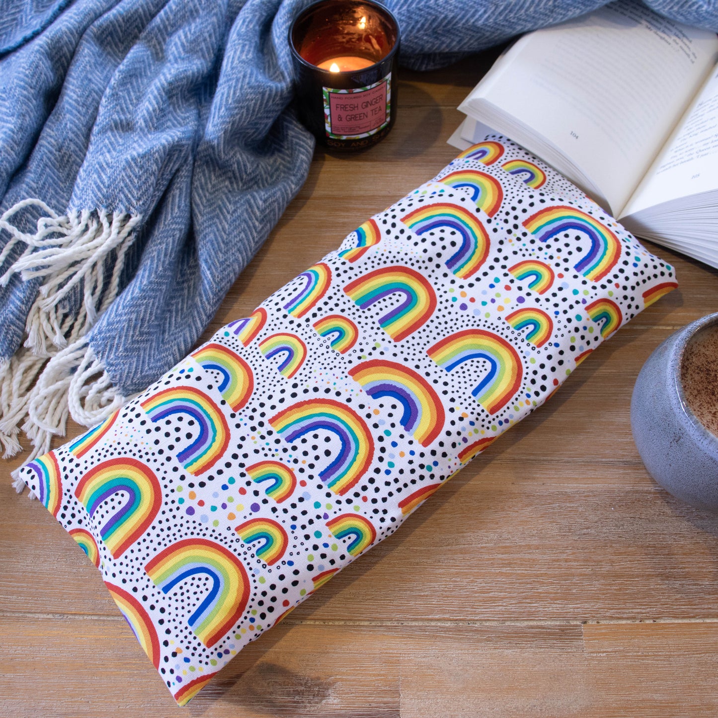 A long rectangular heat pack lies on a wooden surface surrounded by a blue blanket, open book, burning candle and filled coffee cup.  The Heat Pack has a white base and is covered in bright stylised rainbows in various sizes.  Between the rainbows are colourful dots.