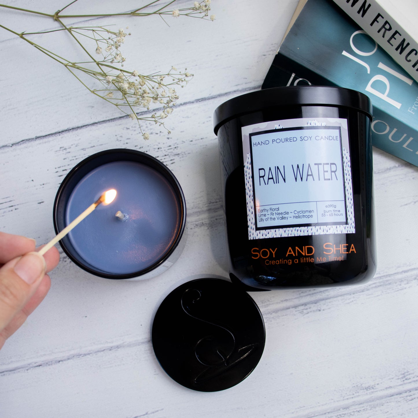 A flat lay photo showing a large black jar lying on its side on a white wooden surface next to a stack of books and a sprig of baby's breath.  To the left of the jar is a smaller candle standing upright and filled with dark blue wax.  A hand reaches in with a lit match.