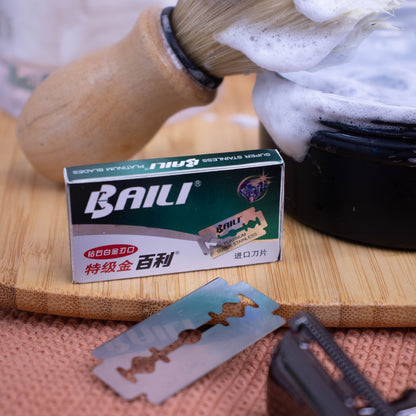 A green and white box of Stainless Steel Razor Blades stands upright on a wooden surface in front of a black tin with dense foam flowing over its side.  In front of the box is a single silver blade.