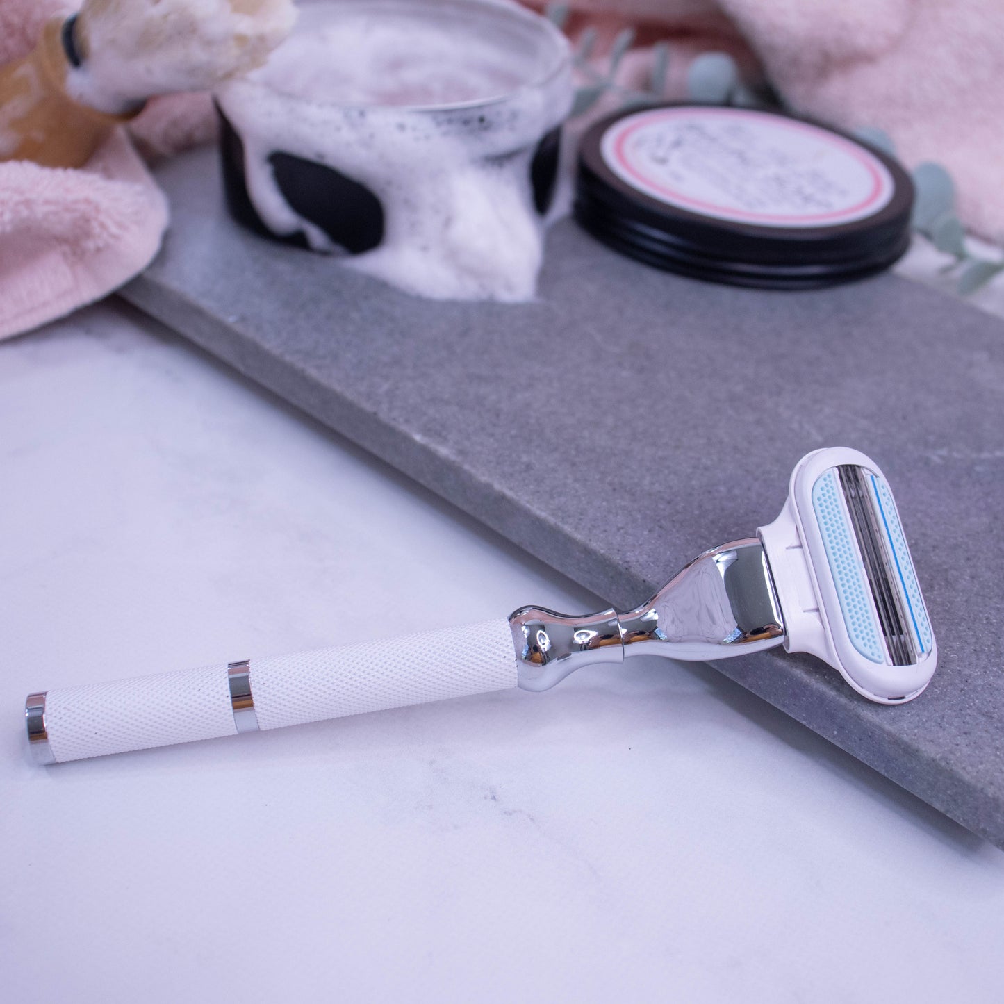 A razor with a textured white handle and silver trim lays across a white surface with the head of the razor, a Venus Cartridge, propped against a grey stone tray.  In the blurred background is a pink towel along with an open tin of shaving soap with a dense lather flowing over the top.