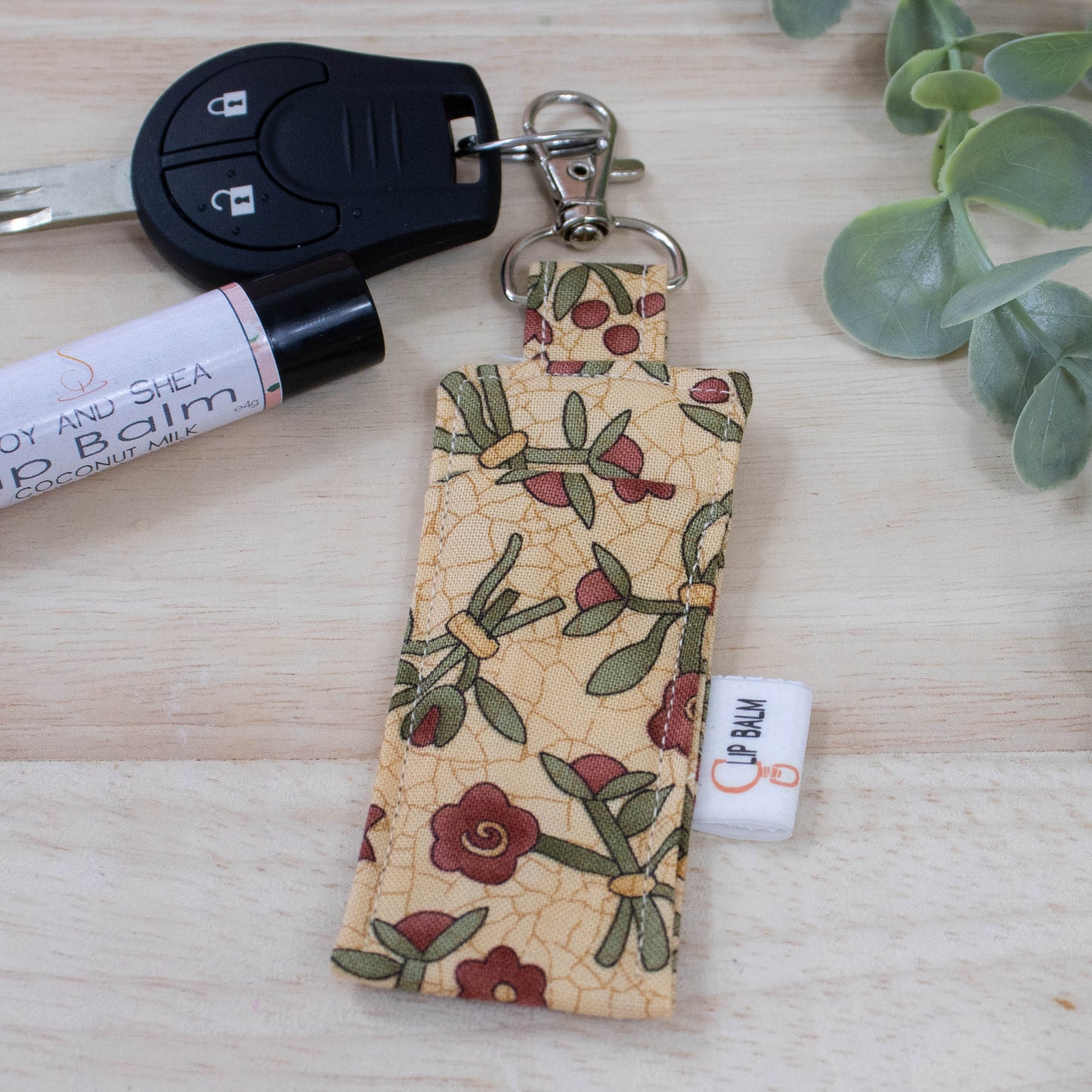 A clip balm holder lies on a wooden surface attached to a car key with a "lobster claw" clip.  To the side is a tube of lip balm and some greenery.  The Clip Balm has a beige background with a crackle pattern in gold and illustrations of flower posies with red flowers and dark green leaves.