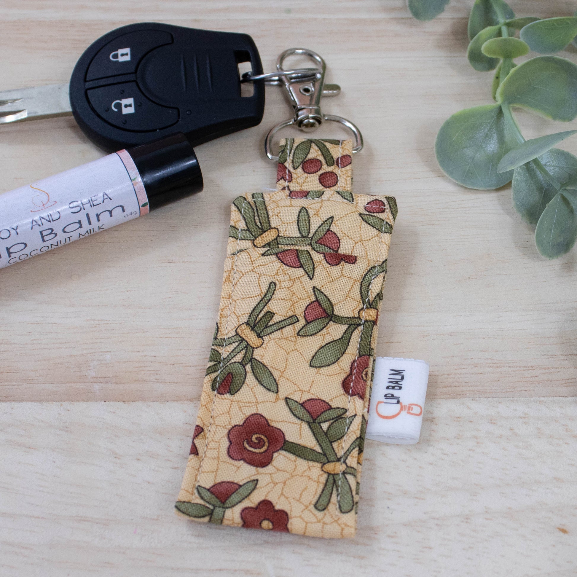 A clip balm holder lies on a wooden surface attached to a car key with a "lobster claw" clip.  To the side is a tube of lip balm and some greenery.  The Clip Balm has a beige background with a crackle pattern in gold and illustrations of flower posies with red flowers and dark green leaves.