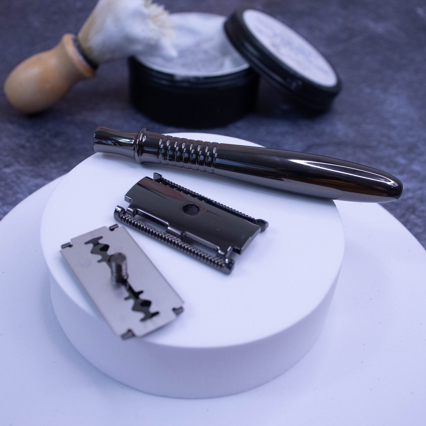 A small white circular foam display sits on a larger one against a dark background with a tin of shaving soap and brush.  Ontop of the small display is a Safety Razor in gunmetal grey that has been dismantled to show a long, roundish handle, the head of the razor and a razor blade.