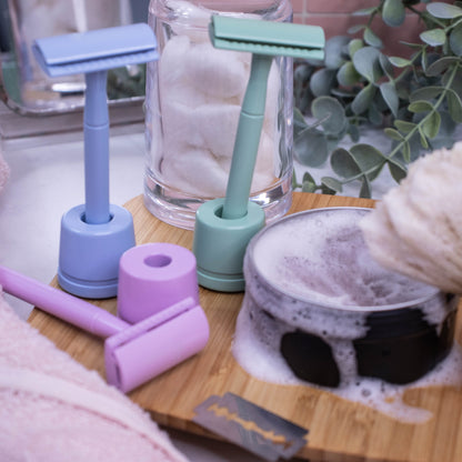 A collection of three safety razors in pale colours of blue, green and pink stand alongside a disposable blade and a tin of shaving soap with a dense lather.  The blue and green razors stand upright in a matching cylindrical stand wher as the pink one lies flat next to its stand. 
